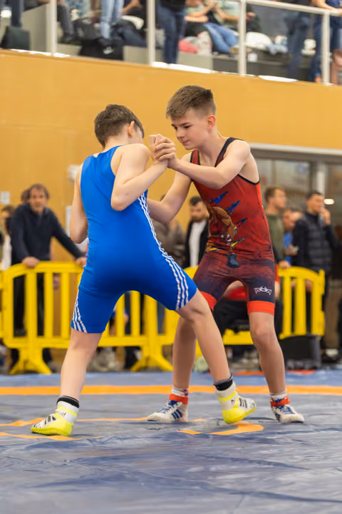 Two young wrestlers grip each other's hands in a tense standoff on the blue mat, spectators watching behind.