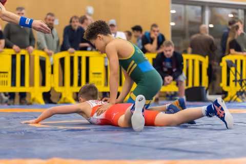 Boy in green singlet dominates opponent in red, pinning him to the blue mat during a youth wrestling bout.