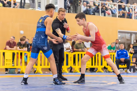 Two young wrestlers in blue and red singlets square off tensely as a referee watches closely in a busy arena.