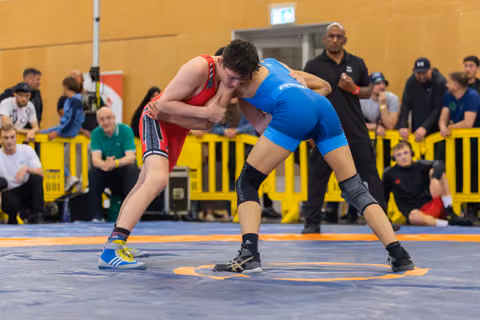 Two young wrestlers lock up in an intense clinch on the mat, red vs blue, surrounded by spectators in an indoor venue.