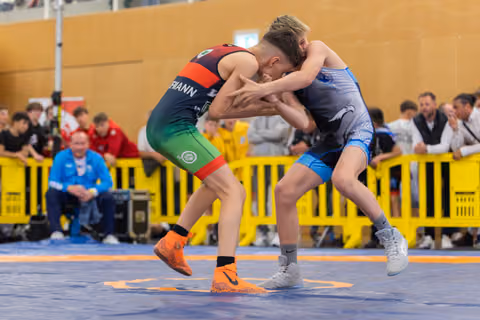 Two youth wrestlers lock in a fierce clinch on the blue mat, one lifted off his feet during an indoor competition.