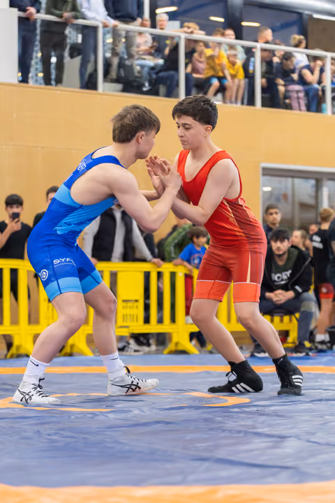 Two young wrestlers in blue and red singlets lock hands in a tense standoff on a blue mat, crowd watching.