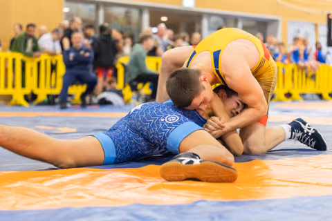Wrestler in blue singlet pinned down by opponent in yellow, both straining intensely on orange and blue mat.