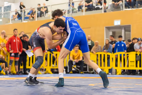 Two wrestlers locked in a fierce grapple on the mat, blue SUI singlet athlete driving forward with determination.