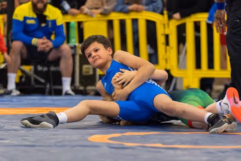 Young boy in blue singlet wrestles opponent to the mat, gripping tightly with fierce concentration during youth bout.