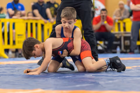 Two boys wrestle on a blue mat; the boy on top focuses intensely, controlling his opponent with a firm grip.