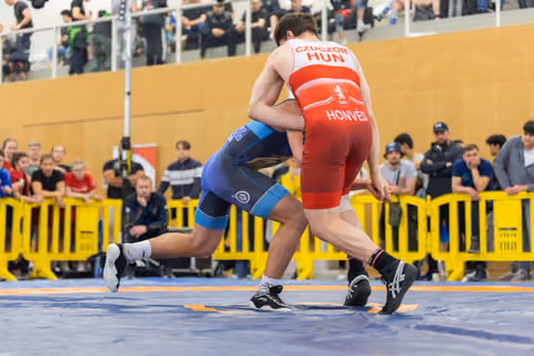 Czuczor (HUN/Honvéd) in red locks up with a blue-suited opponent on the mat, crowds watching behind barriers.