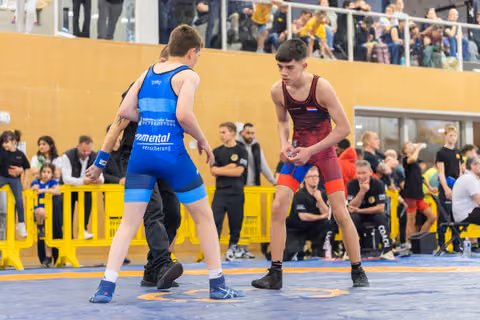 Two teen wrestlers in blue and red singlets square off on the mat, focused and ready to engage at an indoor event.