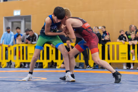 Two young boys in wrestling singlets grip each other intensely on a blue mat, surrounded by spectators behind yellow barriers.