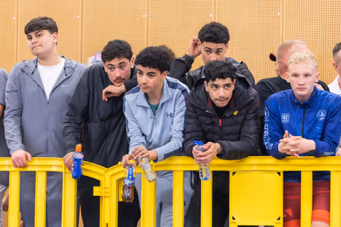 Group of teenage athletes lean on a yellow barrier, watching intently with focused, serious expressions at an indoor sports event.