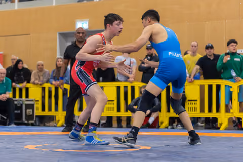 Teen wrestler in red singlet squares off against opponent in blue Phantom singlet on a competition mat, crowd watching.