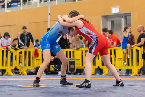 Two young female wrestlers locked in a tight clinch, arms wrapped around each other's necks, intense focus on the mat.