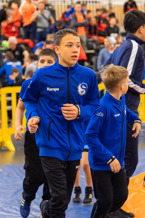 Focused teenage boy in blue Kempa jacket leads younger teammates across the mat at an indoor sports event.
