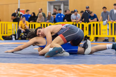 A wrestler in dark singlet pins his opponent to the mat, grimacing with effort, spectators watching behind yellow barriers.