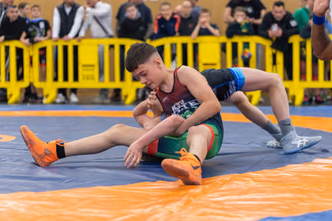 Focused young wrestler controls his opponent on the orange mat, bearing down with intensity before a crowd of spectators.