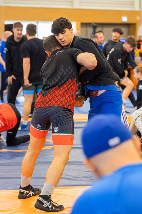 Two wrestlers lock in a tight clinch on an orange mat, surrounded by other athletes training in a busy sports hall.