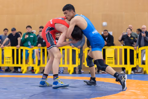 Two wrestlers locked in a fierce grip on the mat, blue singlet athlete dominating red opponent before a crowd.