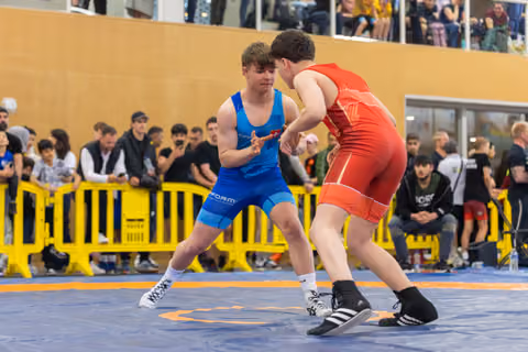 Two teenage wrestlers circle each other on the mat, focused and tense, as a crowd watches from behind yellow barriers.