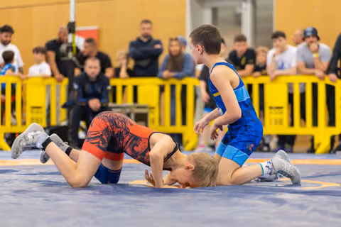 Two boys grapple on a blue mat; one in red is pinned down while the blue-suited boy stands over him, focused.