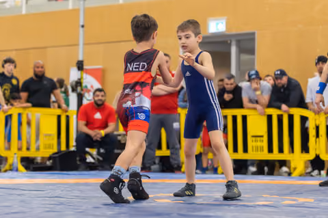 Two young boys in singlets square off on a blue mat, one wearing NED colors, at an indoor youth wrestling event.