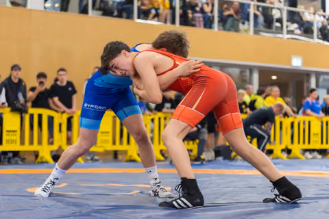 Two young wrestlers lock up on the mat, both focused and determined, competing in blue and red singlets at an indoor event.