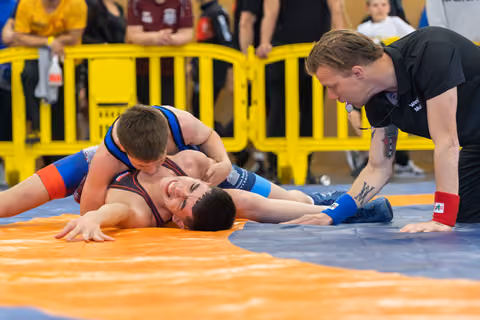A wrestler pins his opponent to the orange mat, grinning, as a tattooed referee leans in closely to watch.