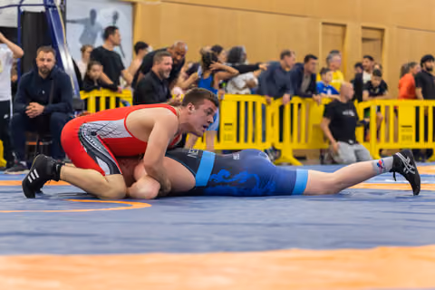 Wrestler in red singlet controls his opponent on the blue mat, leaning forward with focus during an indoor sports event.