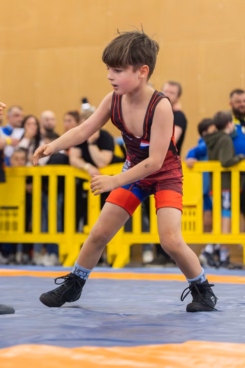 Young boy in red wrestling singlet crouches in a defensive stance on the mat, focused and alert during a match.