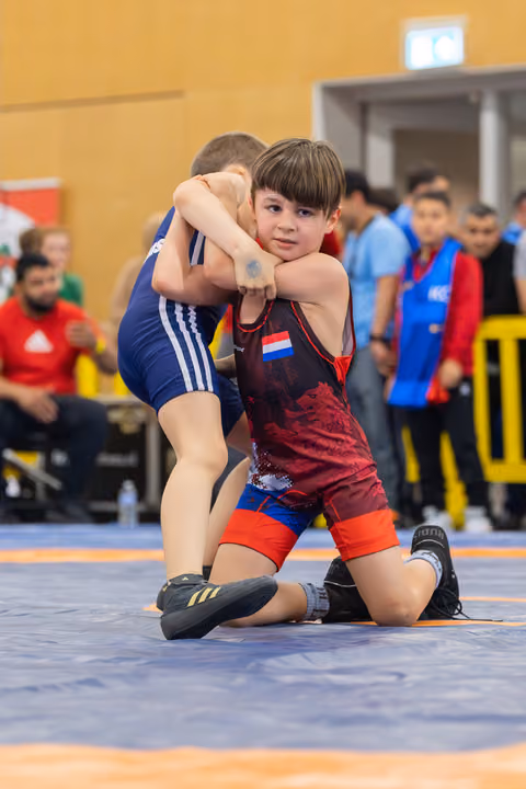 Young Dutch wrestler in red singlet locks opponent's arms, focused expression, kneeling on blue mat at indoor youth event.