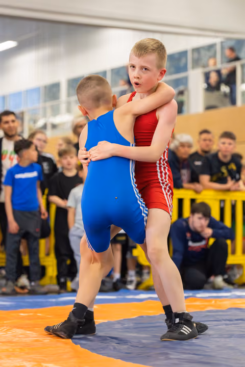 Two young boys in red and blue singlets locked in a tight clinch on the mat, crowd watching intently behind them.