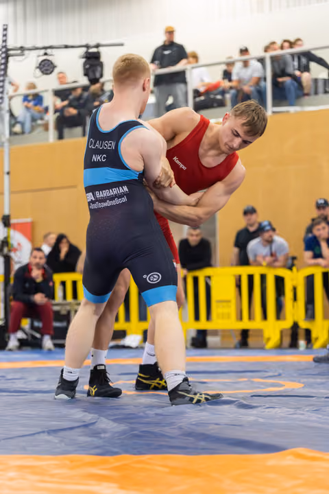 Wrestler Clausen in blue singlet locks arms with red-clad opponent in intense clinch on blue mat, crowd watching