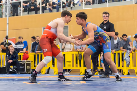 Two young wrestlers lock hands in an intense grip battle, focused and poised on a blue mat before a crowd.