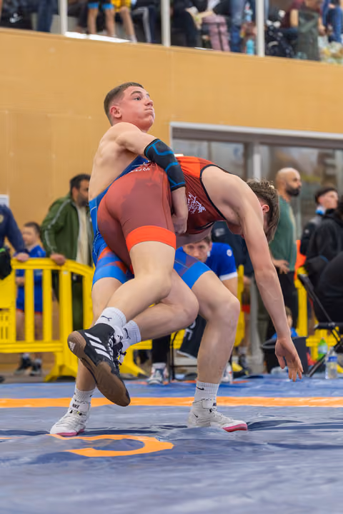 Young wrestler in red singlet lifts opponent off the mat, muscles straining, crowd watching from the stands behind.