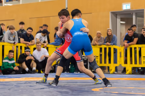 Two young wrestlers lock up intensely on a blue mat, one in red USA singlet, one in blue Phantom, crowd watching.