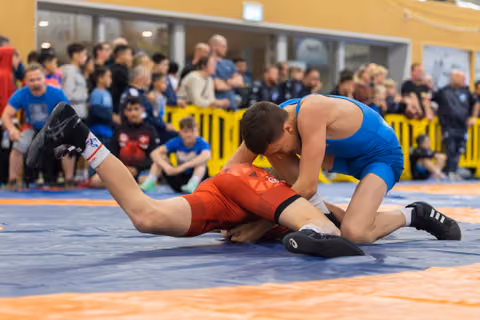 Wrestler in blue singlet dominates opponent in red, pressing him down on the mat while crowd watches intently.