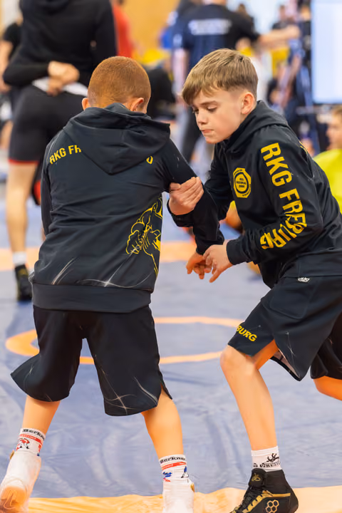 Two focused boys in RKG Freiburg hoodies wrestle intensely on a blue mat, gripping each other's arms.