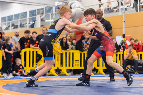 Two young boys face off on the wrestling mat, gripping each other's arms with focused intensity as a referee watches.