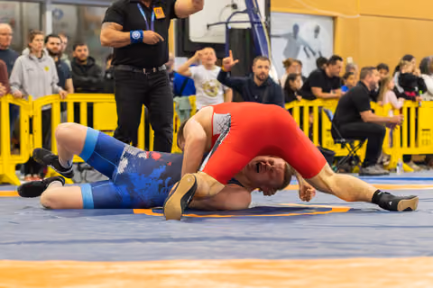 Wrestler in red singlet pins opponent in blue to the mat, crowd reacting with excitement in the background.