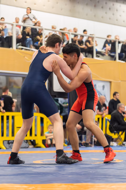 Two young wrestlers in navy and red singlets lock up in a tie position on the mat, focused and competitive.