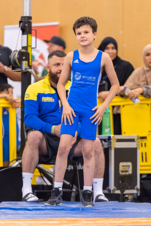 Young boy in blue Adidas singlet stands confidently on the mat, hands on thighs, coach seated behind him.