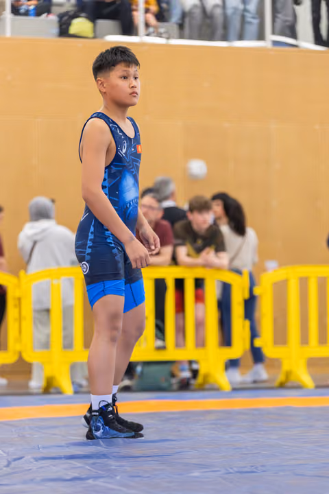 Young wrestler in blue singlet stands alert on the mat, focused expression, awaiting next match at indoor sports hall.