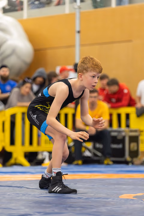 Focused young boy in wrestling singlet crouches in ready stance on blue mat, eyes alert, hands poised.