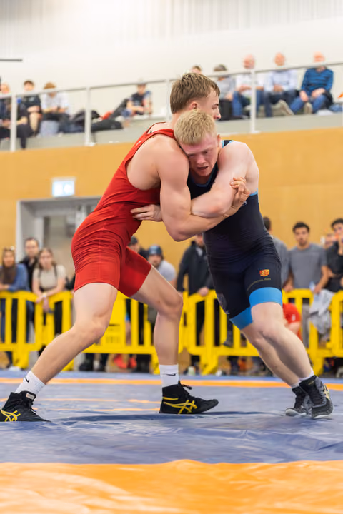 Two young wrestlers locked in a fierce clinch on the mat, both straining with effort, spectators watching from behind yellow barriers.
