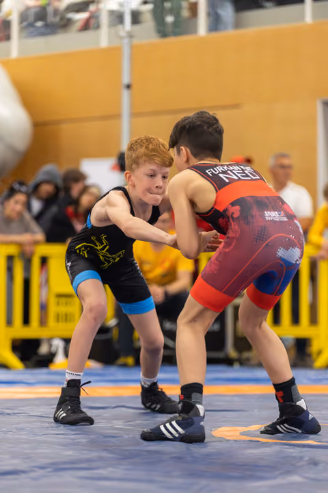 Freckled redhead boy in black singlet squares off against opponent in red NED singlet, both crouched in tense standoff.