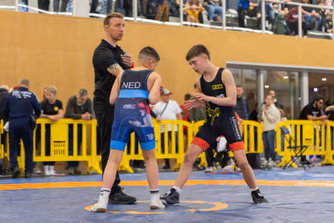 Young wrestler in NED singlet faces opponent in RSK kit as referee steps in to separate them on the blue mat.