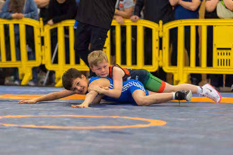 Boy in blue singlet pinned on mat, smiling, as opponent in black and green holds him down at indoor youth match.