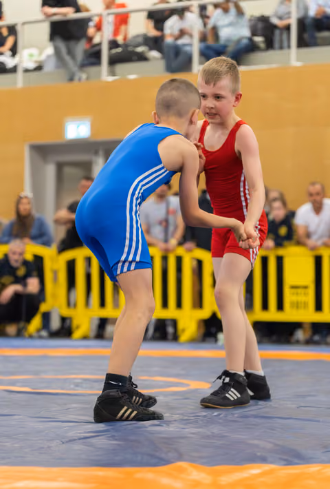 Two young boys in blue and red singlets lock hands in an intense wrestling match on a blue mat, crowd watching.
