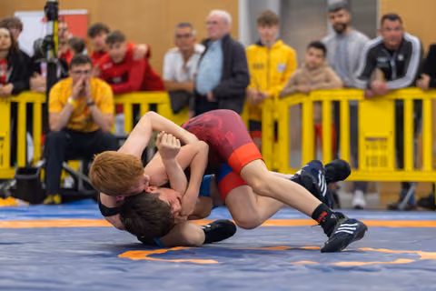 Two young wrestlers locked in a ground hold on the mat, crowd of coaches and spectators watching intently behind yellow barriers.
