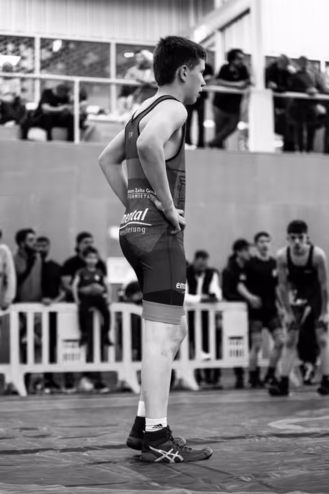 Teen wrestler stands still, hands on hips, composed and focused, watching match from the side of the mat.