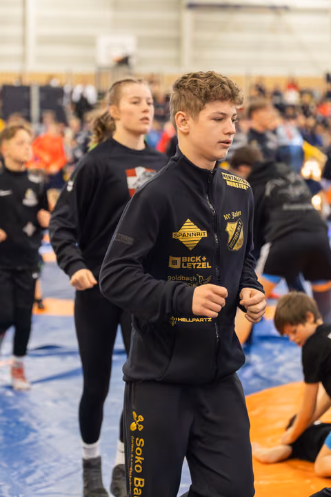 Focused teen wrestler in black sponsor jacket jogs during warm-up in a busy indoor sports hall.